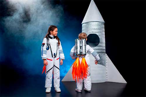 Niños usando **disfraces con material reciclado fáciles** de astronauta con tanques de oxígeno hechos de botellas de plástico plateadas y un cohete de cartón.