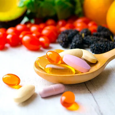Cuchara de madera con cápsulas de suplementos vitamínicos frente a frutas y verduras frescas, destacando la importancia de las vitaminas en el metabolismo.