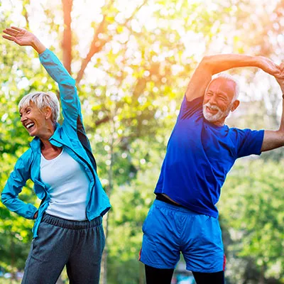 Pareja de adultos mayores sonriendo mientras realizan rutinas de ejercicios de estiramiento al aire libre.