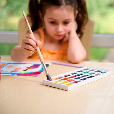 Niña pequeña desarrollando su talento creativo mientras pinta en su mesa con materiales de arte como pincel y acuarelas