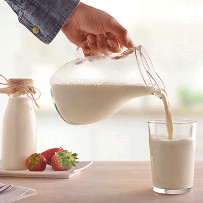Una mano vertiendo leche fresca de una jarra de vidrio en un vaso, con una botella de leche y fresas frescas al fondo sobre una mesa de madera. Imagen que ilustra el consumo de leche animal y sus beneficios nutricionales.