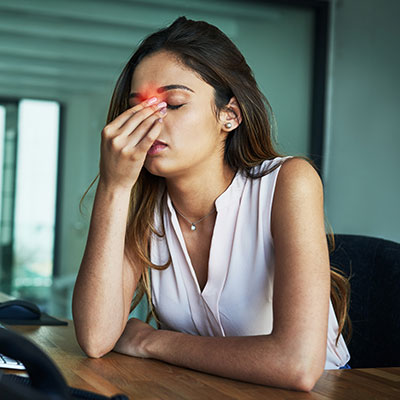 Mujer en una oficina con los ojos cerrados y tocándose el tabique nasal, reflejando agotamiento y síntomas de fatiga crónica.