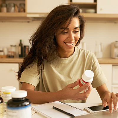 Mujer joven en una cocina revisando la etiqueta de un frasco de suplementos nutricionales mientras usa su laptop para investigar.