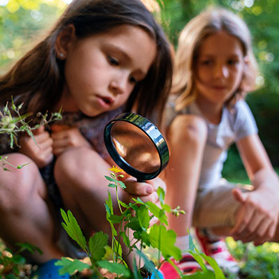 Dos niñas aplicando las etapas de exploración en los niños al observar plantas con una lupa en un jardín.