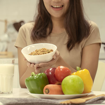 Consejos para una dieta rica en prebióticos. Mujer sonriente sosteniendo un tazón de cereales integrales junto a vegetales frescos, ilustrando las claves para incorporar prebióticos en la alimentación.