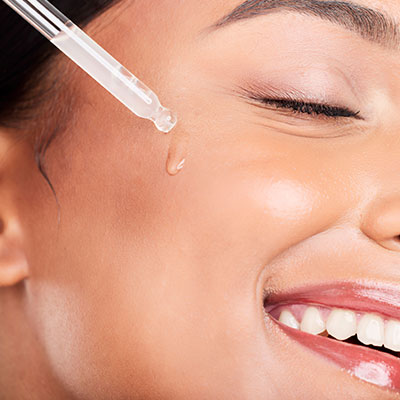 Mujer sonriente aplicando gotas de sérum para aprovechar los beneficios del ácido hialurónico en la piel del rostro.