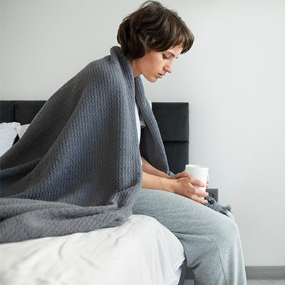Mujer enferma sentada en la cama envuelta en una cobija sosteniendo una taza caliente, experimentando síntomas de defensas bajas.