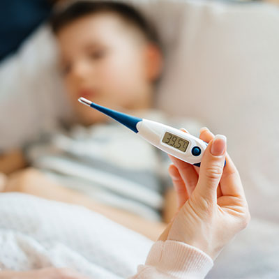Mano sosteniendo un termómetro digital que marca 39,5 °C frente a un niño acostado en cama.
